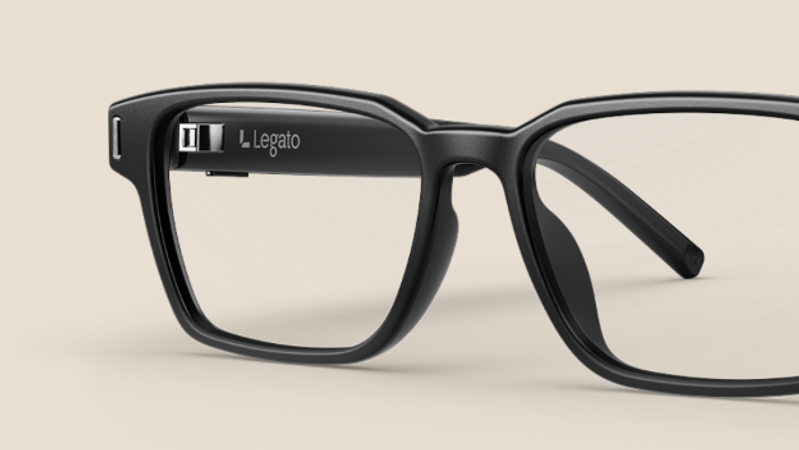 The image showcases a close-up of stylish black eyeglasses with a modern design. The frame features a logo on the inner side, emphasizing its contemporary appeal.