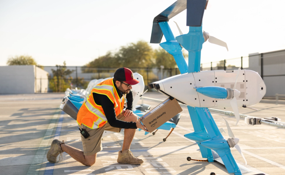A worker in a safety vest kneels beside a blue and white drone, preparing it for flight. The background features several drones lined up on a paved surface, indicating a drone delivery or operation facility.