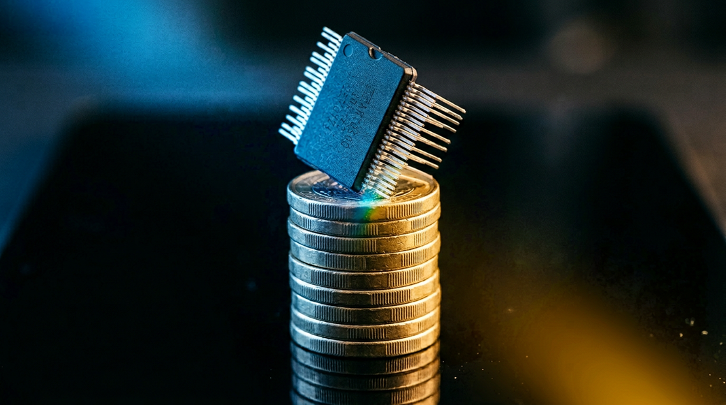An abstract close-up photograph featuring a microchip balanced precariously on its edge atop a vertical stack of British one-pound coins. The cool blue light enhances the chip's matte black surface, while warm gold tones reflect off the edges of the coins, suggesting a juxtaposition of technology and capital in a clean, modern composition.