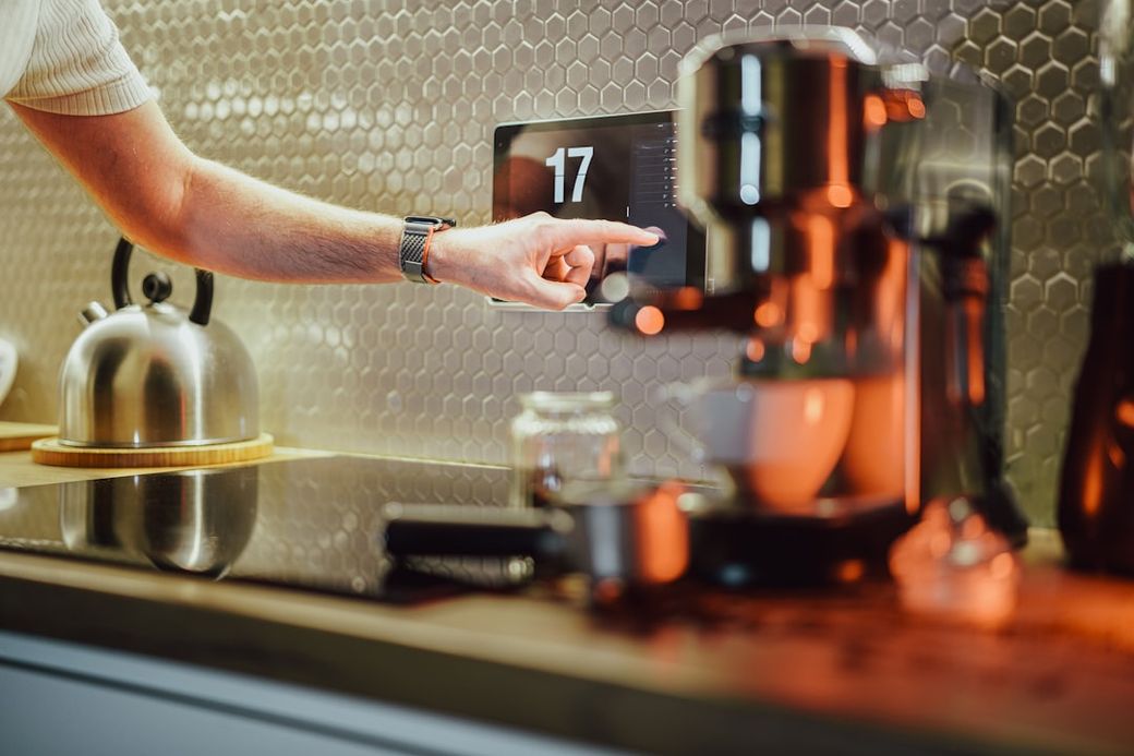 A close-up of a person pointing at a digital display on a coffee machine in a modern kitchen. The setting features sleek design elements, including a kettle in the background and a polished countertop. — Credit: Photo by Jakub Żerdzicki on Unsplash
