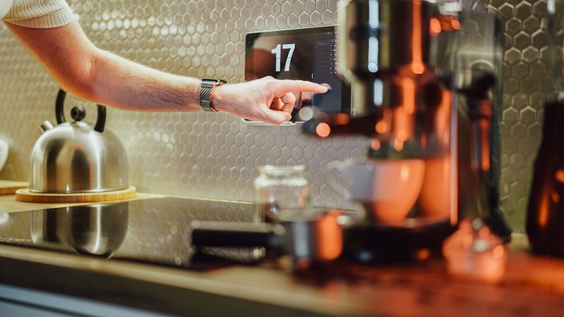 A close-up of a person pointing at a digital display on a coffee machine in a modern kitchen. The setting features sleek design elements, including a kettle in the background and a polished countertop.