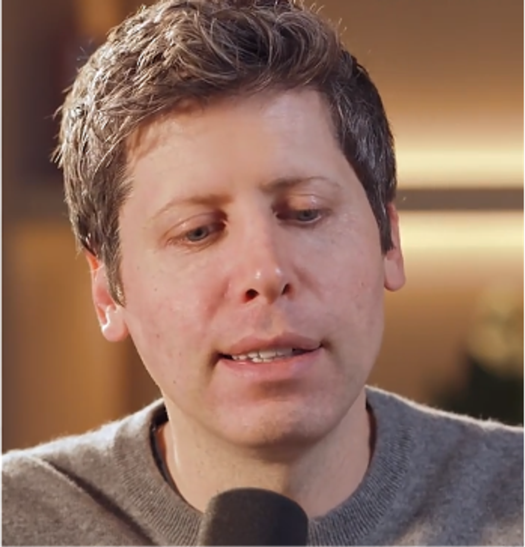 The image features a close-up of a man with short, light brown hair, wearing a gray sweater. He appears to be engaged in conversation, possibly speaking into a microphone, suggesting a setting that may involve a discussion or interview on technology or innovation, themes often associated with Sam Altman.