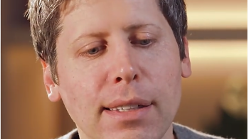 The image features a close-up of a man with short, light brown hair, wearing a gray sweater. He appears to be engaged in conversation, possibly speaking into a microphone, suggesting a setting that may involve a discussion or interview on technology or innovation, themes often associated with Sam Altman.