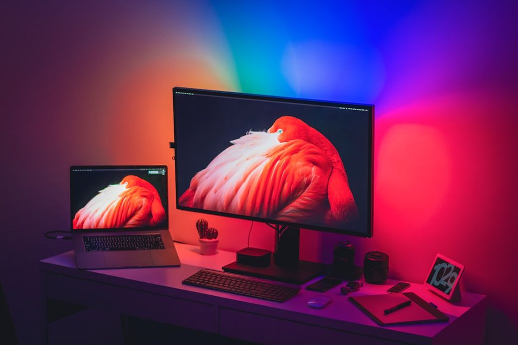 The image features a modern workspace with two screens displaying vibrant pink and orange hues. The setup includes a laptop and a larger monitor, illuminated by multicolored ambient lighting. — Credit: Photo by Virul Weerasooriya on Unsplash