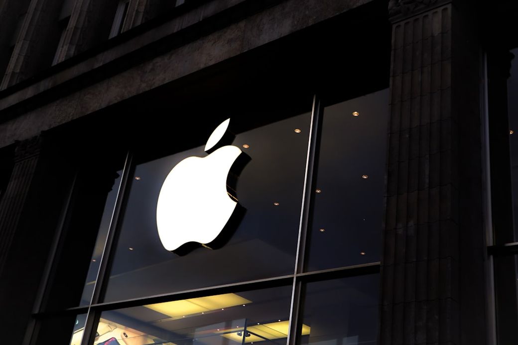 An exterior view of an Apple retail store featuring a large illuminated Apple logo. The image captures the store's glass entrance and modern architectural design. — Credit: Photo by Laurenz Heymann on Unsplash