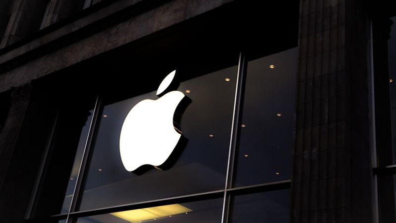 An exterior view of an Apple retail store featuring a large illuminated Apple logo. The image captures the store's glass entrance and modern architectural design.