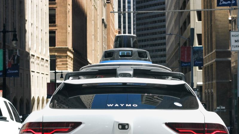 The image features a Waymo autonomous vehicle driving through a city street, with tall buildings lining the sides. The vehicle is equipped with sensors and cameras on its roof, indicating its self-driving capabilities.