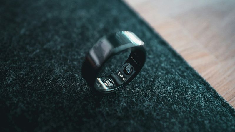 The image features a close-up of a sleek black ring placed on a textured dark surface, highlighting its polished design. The background shows hints of wood flooring, providing a contrast to the ring's dark color.
