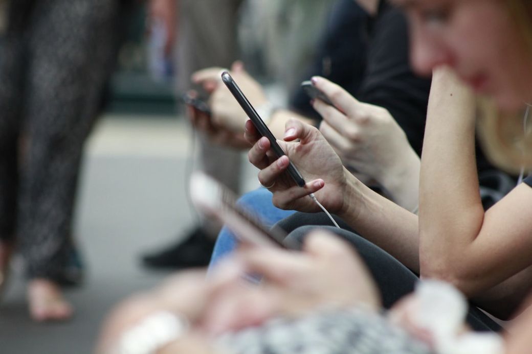 The image captures a group of individuals sitting closely together while focused on their smartphones. Many people are seen with earbuds, suggesting they are engaged in personal activities such as messaging or listening to music. — Credit: Photo by ROBIN WORRALL on Unsplash
