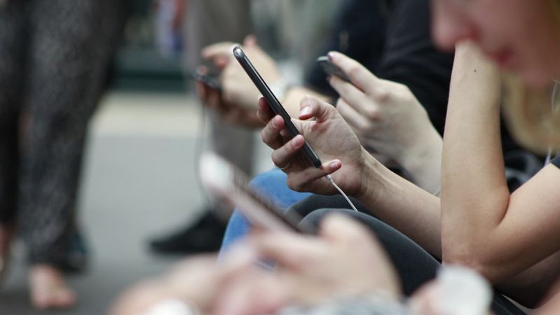 The image captures a group of individuals sitting closely together while focused on their smartphones. Many people are seen with earbuds, suggesting they are engaged in personal activities such as messaging or listening to music.