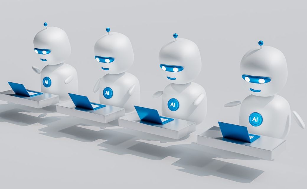 The image features four stylized robots sitting at desks with laptops. Each robot has an illuminated 'AI' symbol on their chest and is engaging with the laptops in front of them, suggesting a focus on artificial intelligence and technology. — Credit: Photo by Mohamed Nohassi on Unsplash