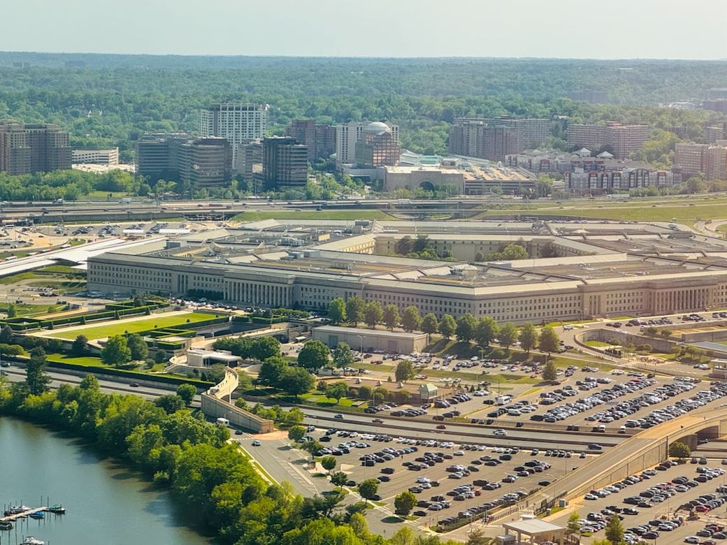 An aerial view of the Pentagon, the headquarters of the United States Department of Defense, surrounded by urban development and green landscapes. The image showcases the building's distinctive five-sided shape and the extensive parking areas nearby. — Credit: Photo by Kevin Doyle on Unsplash