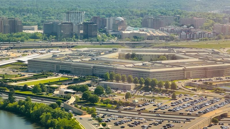 An aerial view of the Pentagon, the headquarters of the United States Department of Defense, surrounded by urban development and green landscapes. The image showcases the building's distinctive five-sided shape and the extensive parking areas nearby.