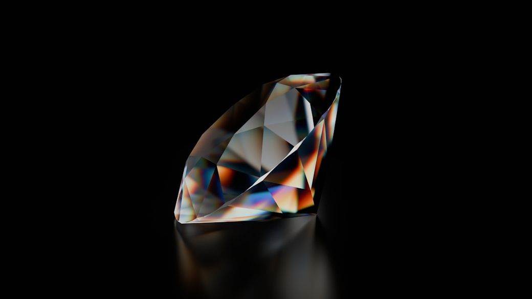 The image features a large, multifaceted diamond resting on a reflective surface. The gem displays a spectrum of colors due to light refraction, set against a black background, emphasizing its cut and brilliance. — Credit: Photo by A Chosen Soul on Unsplash