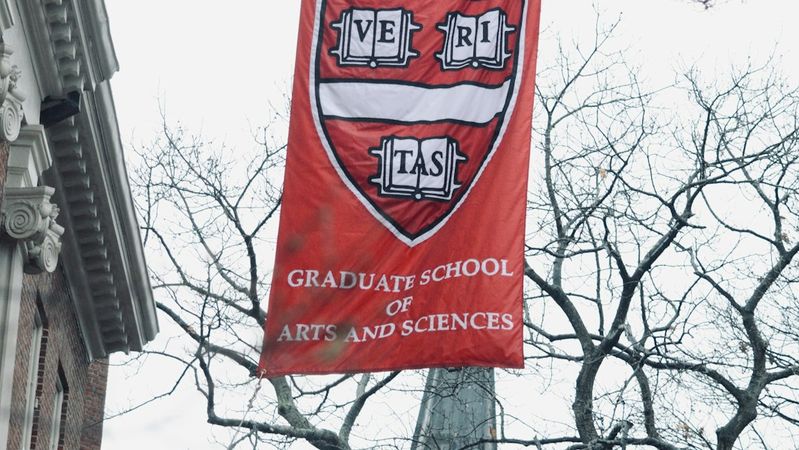 A banner for Harvard University hangs prominently from a building, showcasing the university's crest. The banner also indicates the Graduate School of Arts and Sciences.