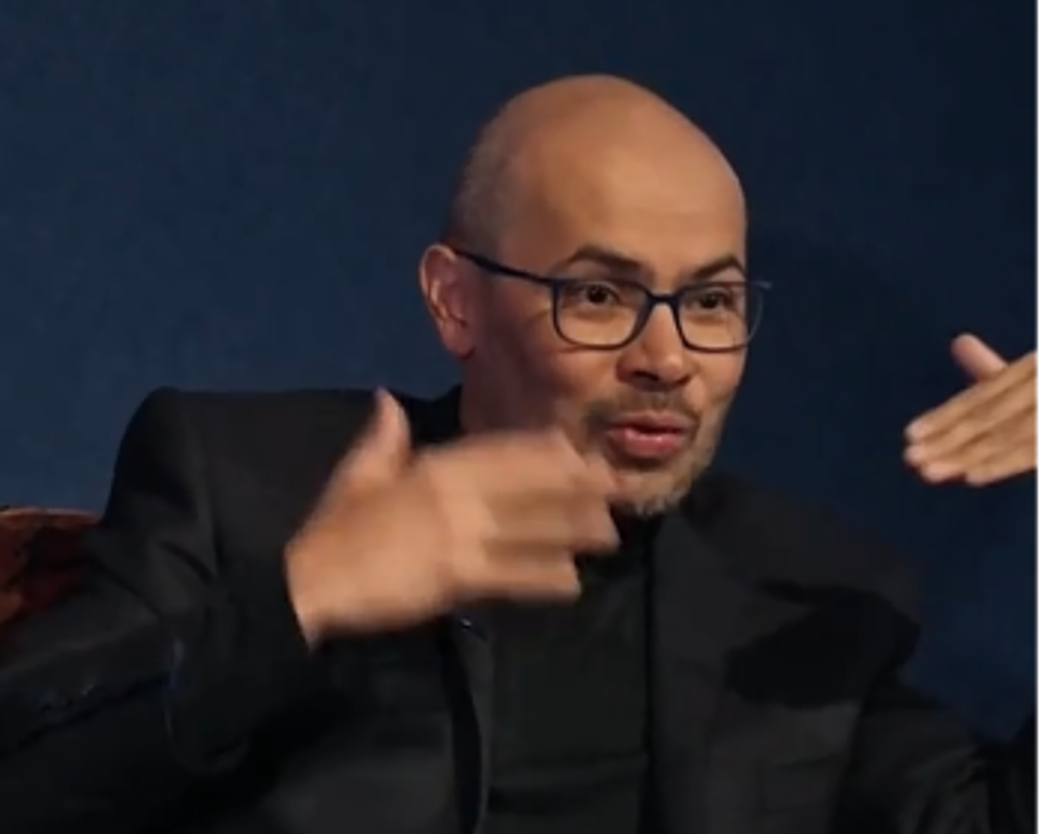 A well-dressed man is engaged in a discussion, using hand gestures to emphasize his points. His expression indicates thoughtful consideration, and he appears to be in a formal interview or panel setting, suggesting a focus on intellectual discourse.