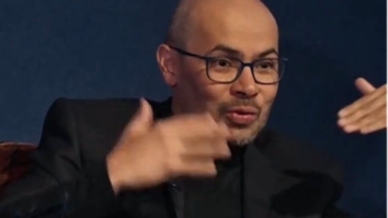 A well-dressed man is engaged in a discussion, using hand gestures to emphasize his points. His expression indicates thoughtful consideration, and he appears to be in a formal interview or panel setting, suggesting a focus on intellectual discourse.