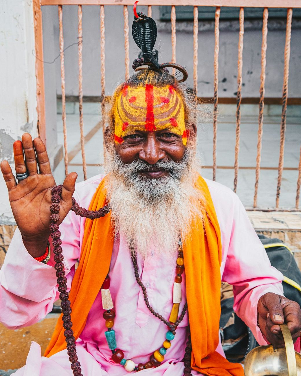 The image features a smiling sadhu, or Hindu holy man, with a colorful orange and yellow painted forehead. He is wearing traditional attire, including a pink garment and orange shawl, and is adorned with beads and other accessories. — Credit: Photo by Jannes Jacobs on Unsplash