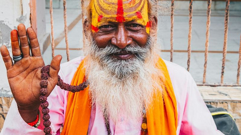 The image features a smiling sadhu, or Hindu holy man, with a colorful orange and yellow painted forehead. He is wearing traditional attire, including a pink garment and orange shawl, and is adorned with beads and other accessories.