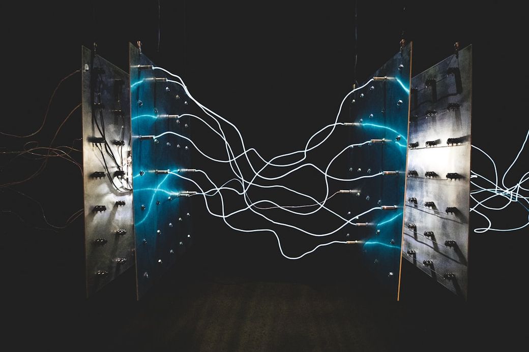 The image displays an abstract arrangement of metallic panels connected by glowing wires, creating a visually striking composition. The contrasting lights against a dark backdrop evoke a sense of technological innovation. — Credit: Photo by israel palacio on Unsplash