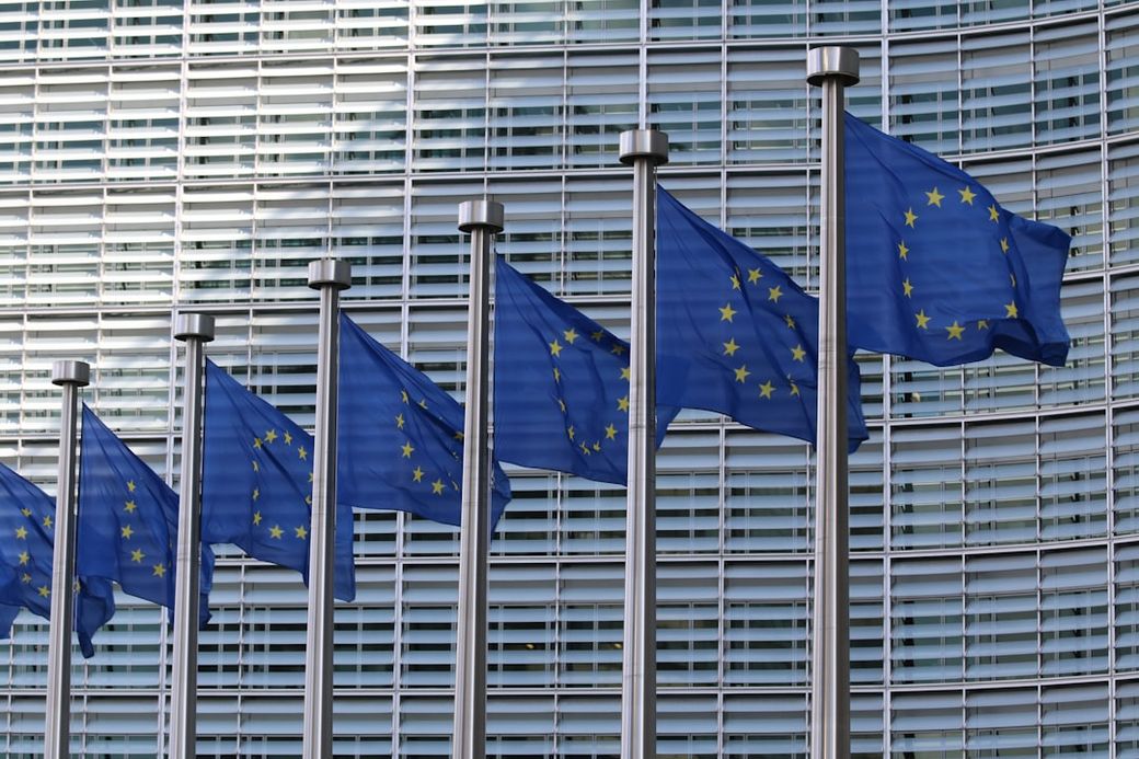 The image features the flags of the European Union displayed prominently outside a modern building. The flags are arranged in a row, fluttering in the breeze against a backdrop of contemporary architecture. — Credit: Photo by Guillaume Périgois on Unsplash