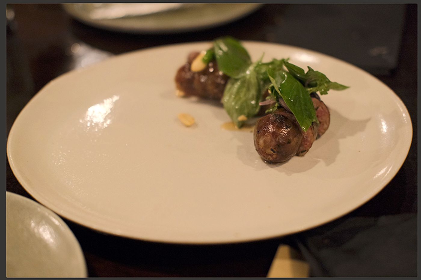 Grilled sausage-like morsels with a tuft of fresh herbs and scattered nuts on a wide white plate