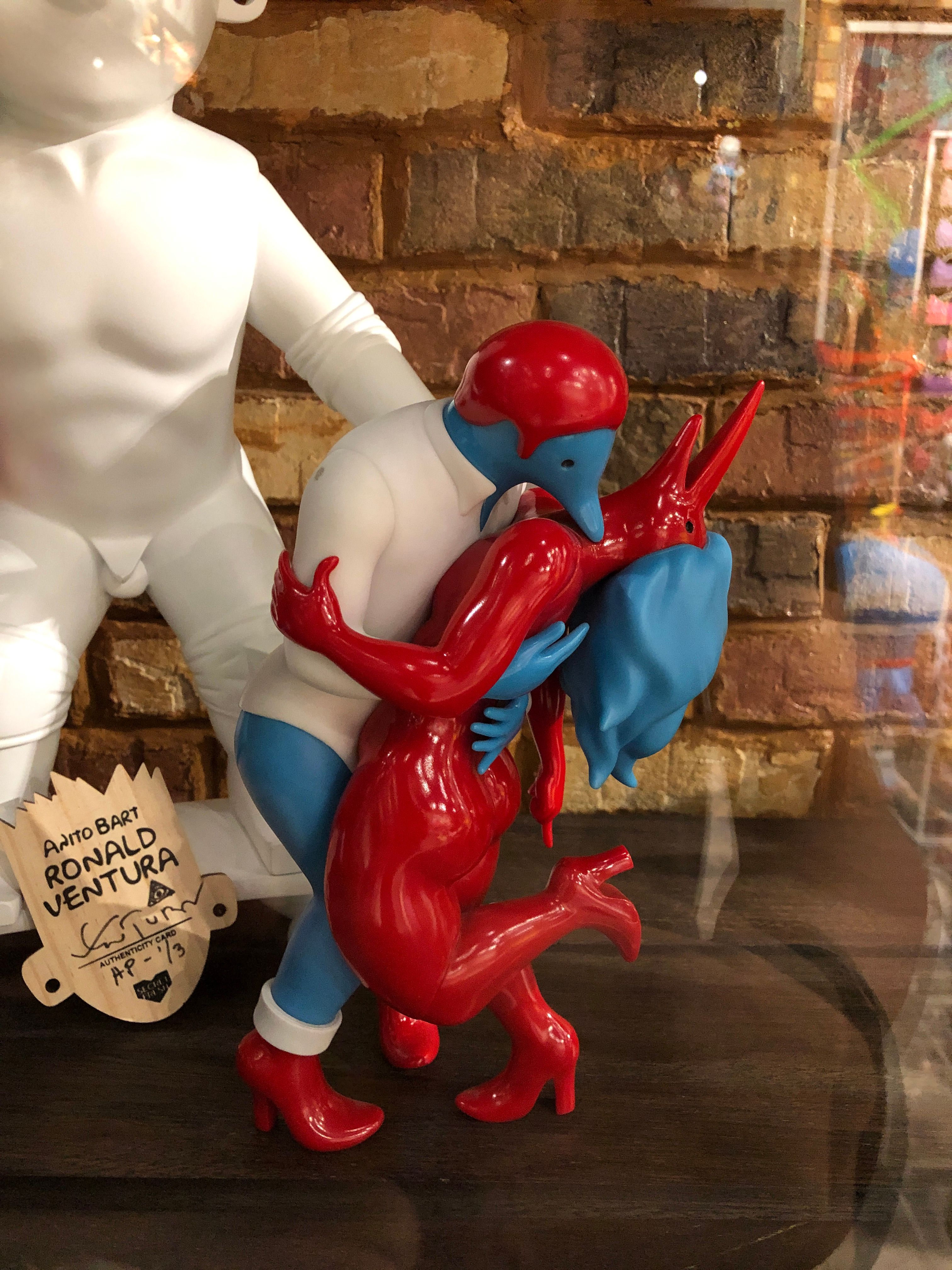 Ronald Ventura’s Anito Bart art toy in a vitrine, a red horned figure in high heels clasped to a blue and white Bart Simpson, its signed authenticity tag beside it