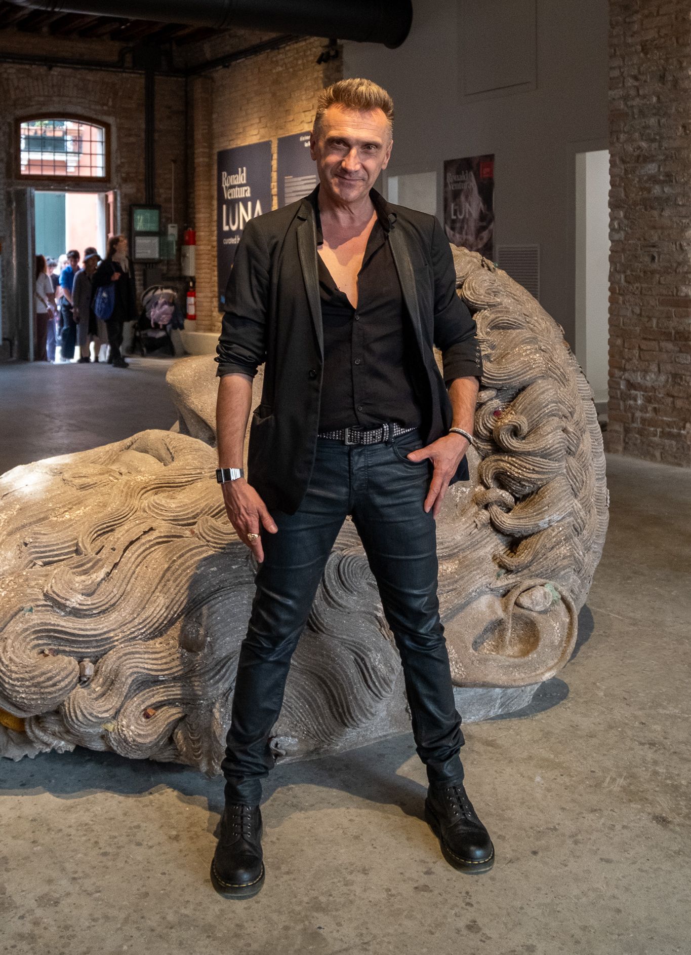 Stefano Cagol in a dark jacket poses before the giant swirling-haired head sculpture in the “Luna” hall
