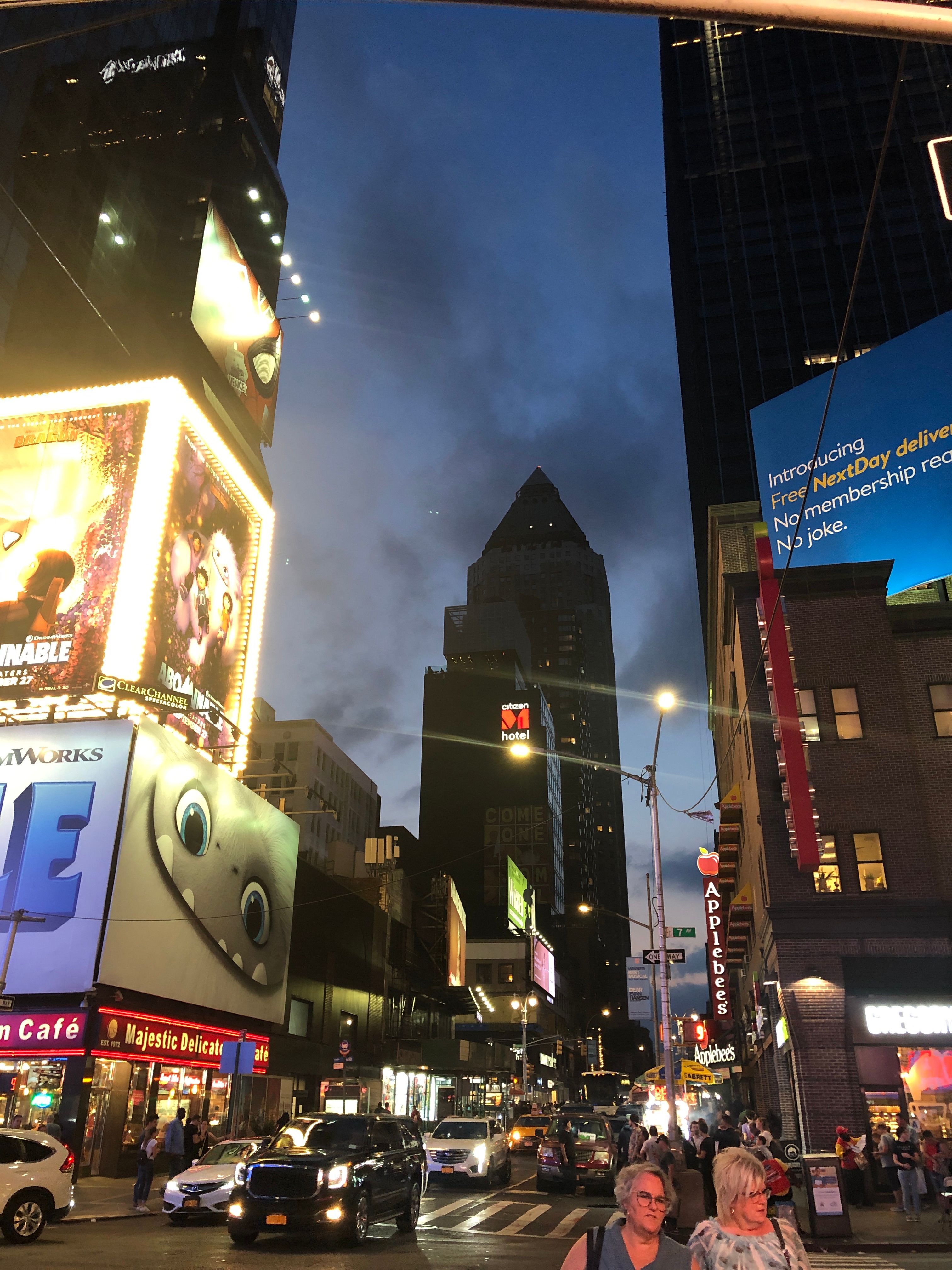 Evening falls on a busy Manhattan street lined with glowing billboards, delis and yellow-lit marquees