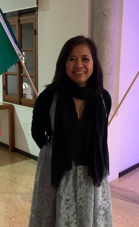 A smiling woman in a black scarf and cardigan and a grey lace skirt stands in a columned hall beside a green flag