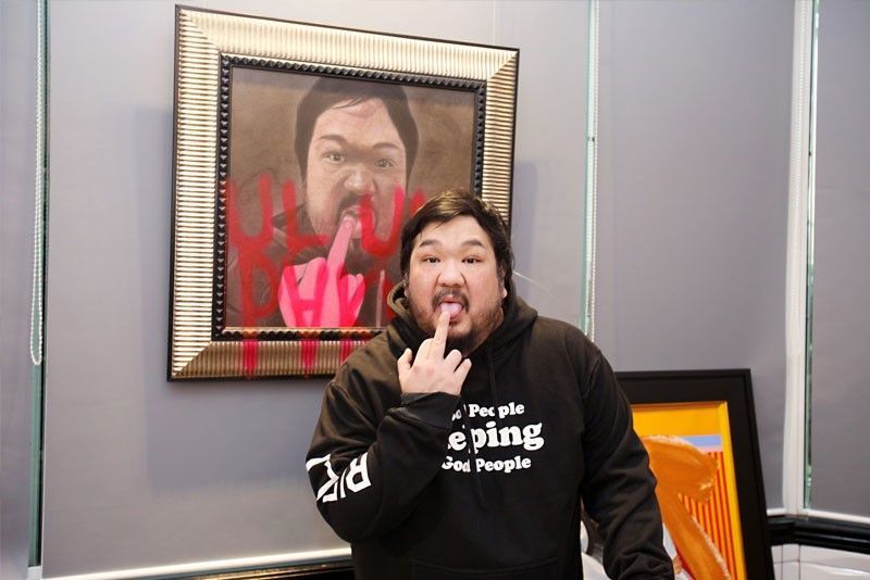 Bigboy Cheng in a black hoodie holds a finger to his lips in front of a framed portrait of himself defaced with red spray paint