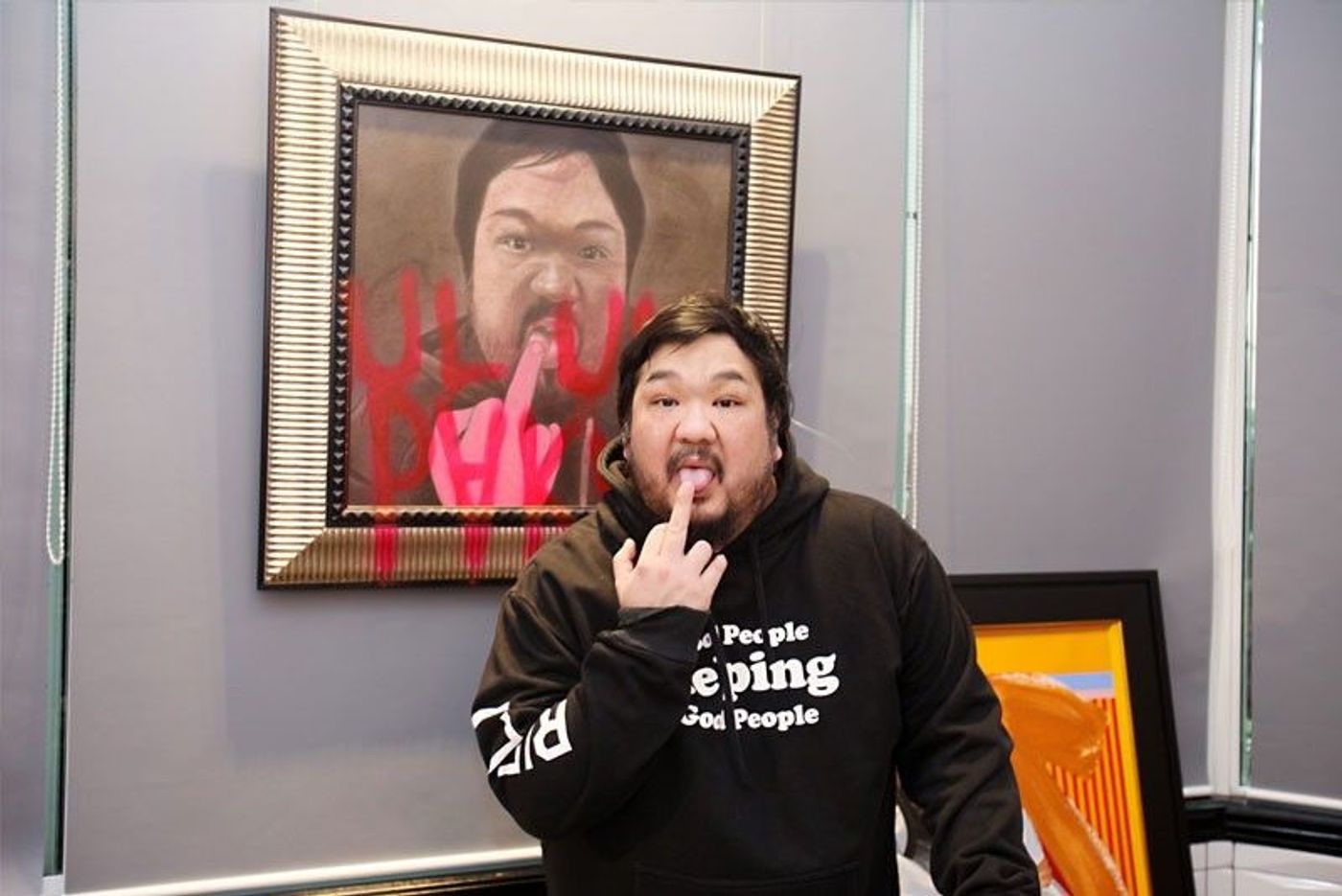 Bigboy Cheng in a black hoodie holds a finger to his lips in front of a framed portrait of himself defaced with red spray paint