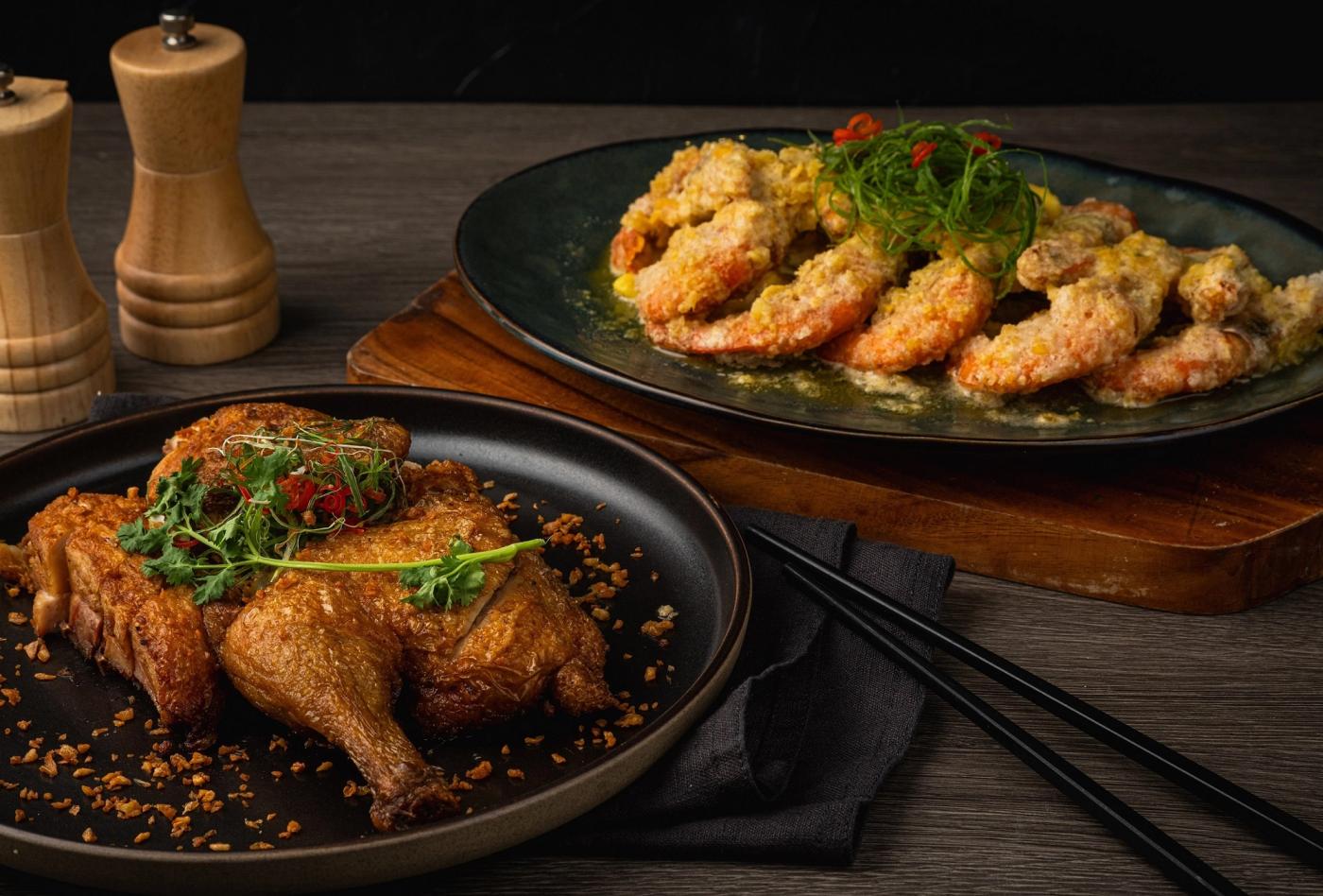 Two Red Lotus dishes on a dark table: a crisp-skinned roast duck and shrimp in golden salted-egg-yolk sauce