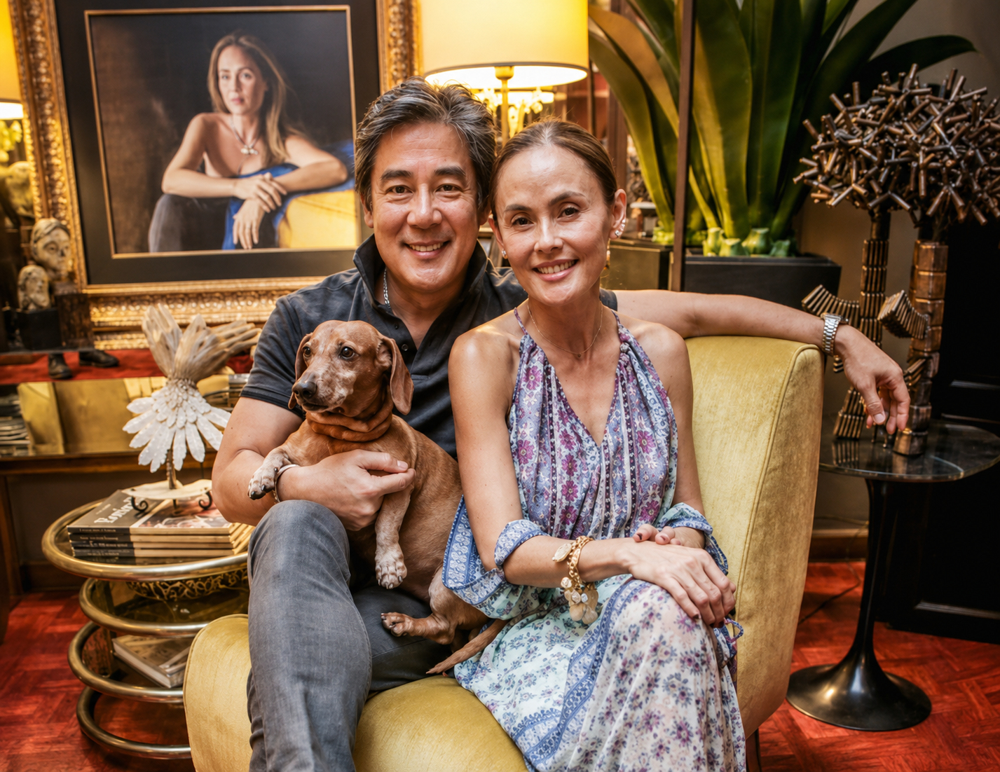 Raul Francisco and Joanna Preysler-Francisco sit close on a mustard sofa holding their brown dachshund, a framed portrait and gilded lamp behind them