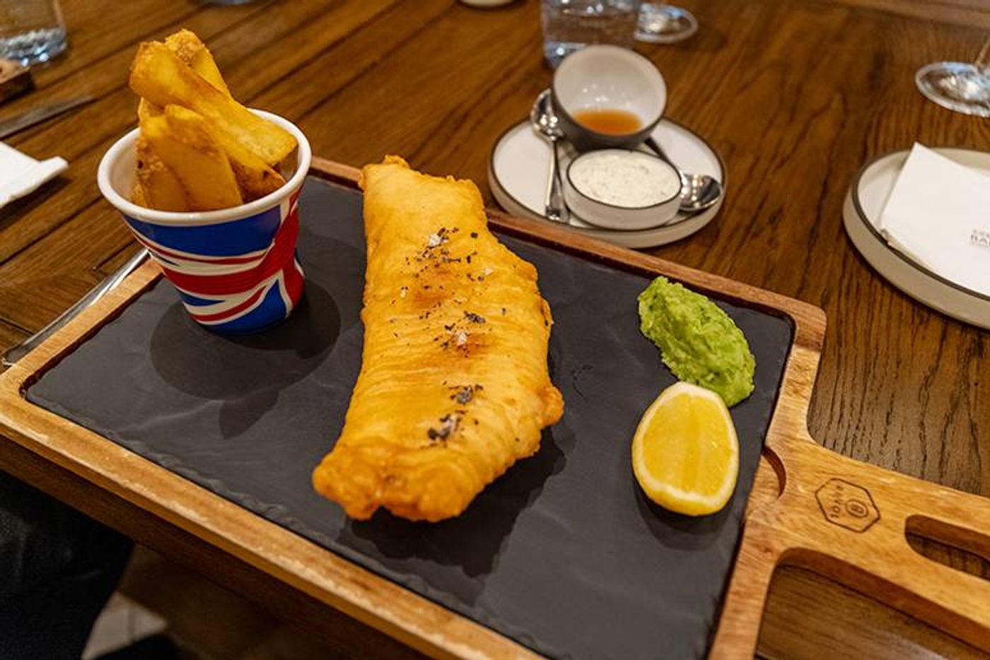 Golden battered fish on a slate board with chips in a Union Jack cup, mushy peas and lemon