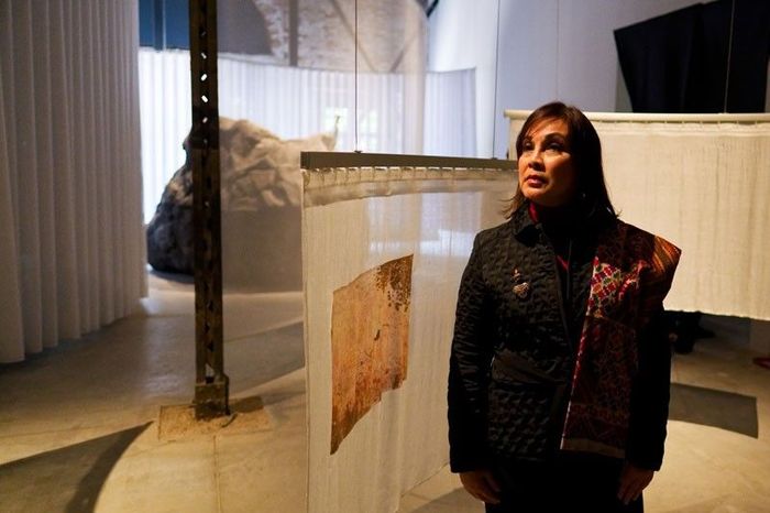 Senator Loren Legarda in a woven-trimmed black jacket stands among hanging fabric artworks and a boulder in the Philippine Pavilion