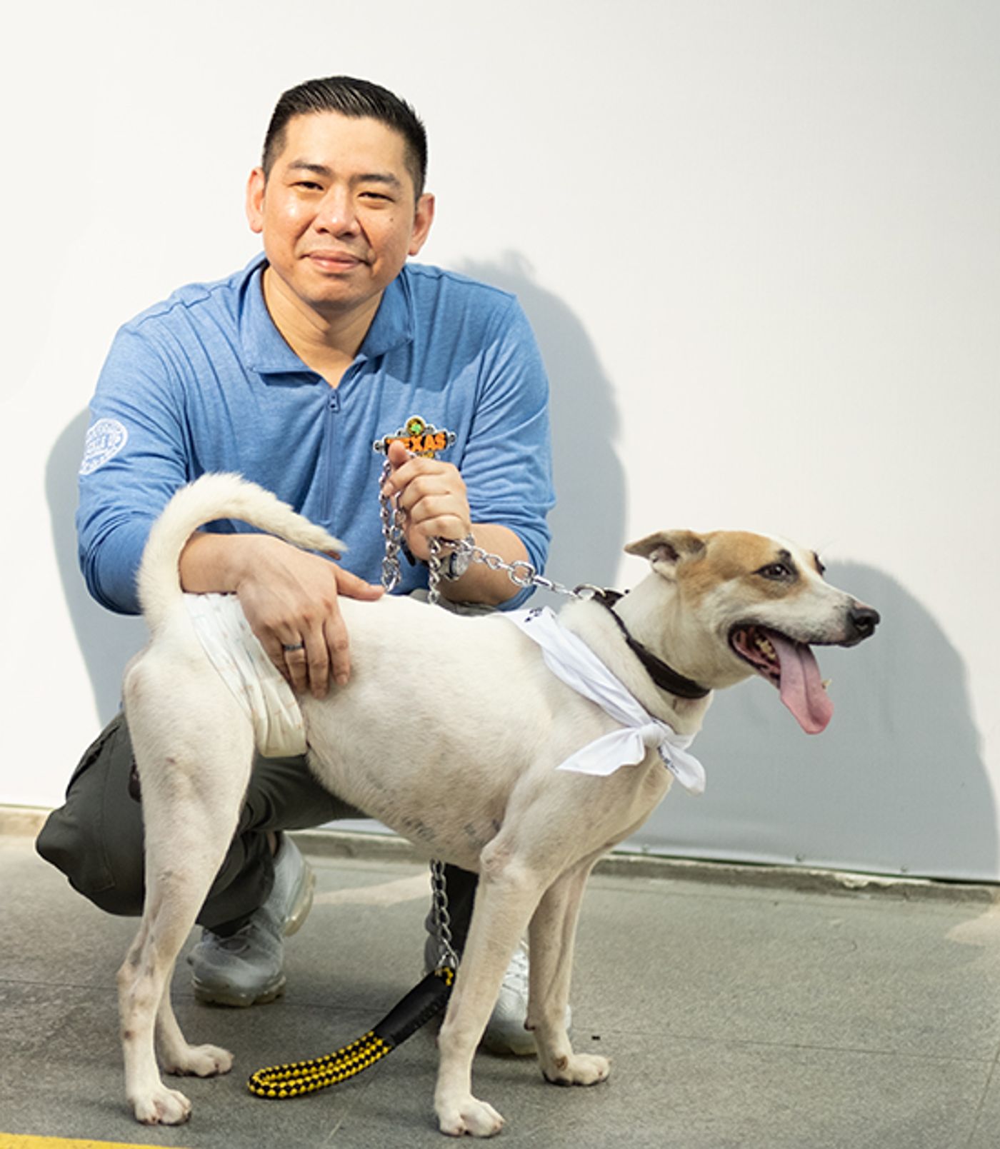 Cyril Reyes, in a blue polo, crouches holding the leash of Texas, a lean white-and-tan dog with its tongue out