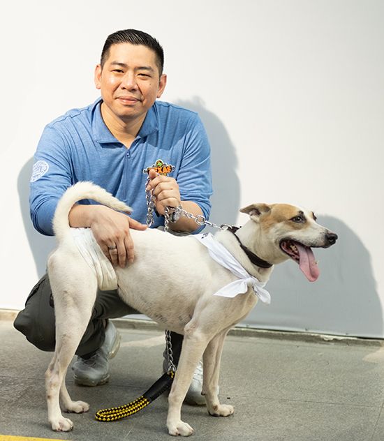 Cyril Reyes, in a blue polo, crouches holding the leash of Texas, a lean white-and-tan dog with its tongue out