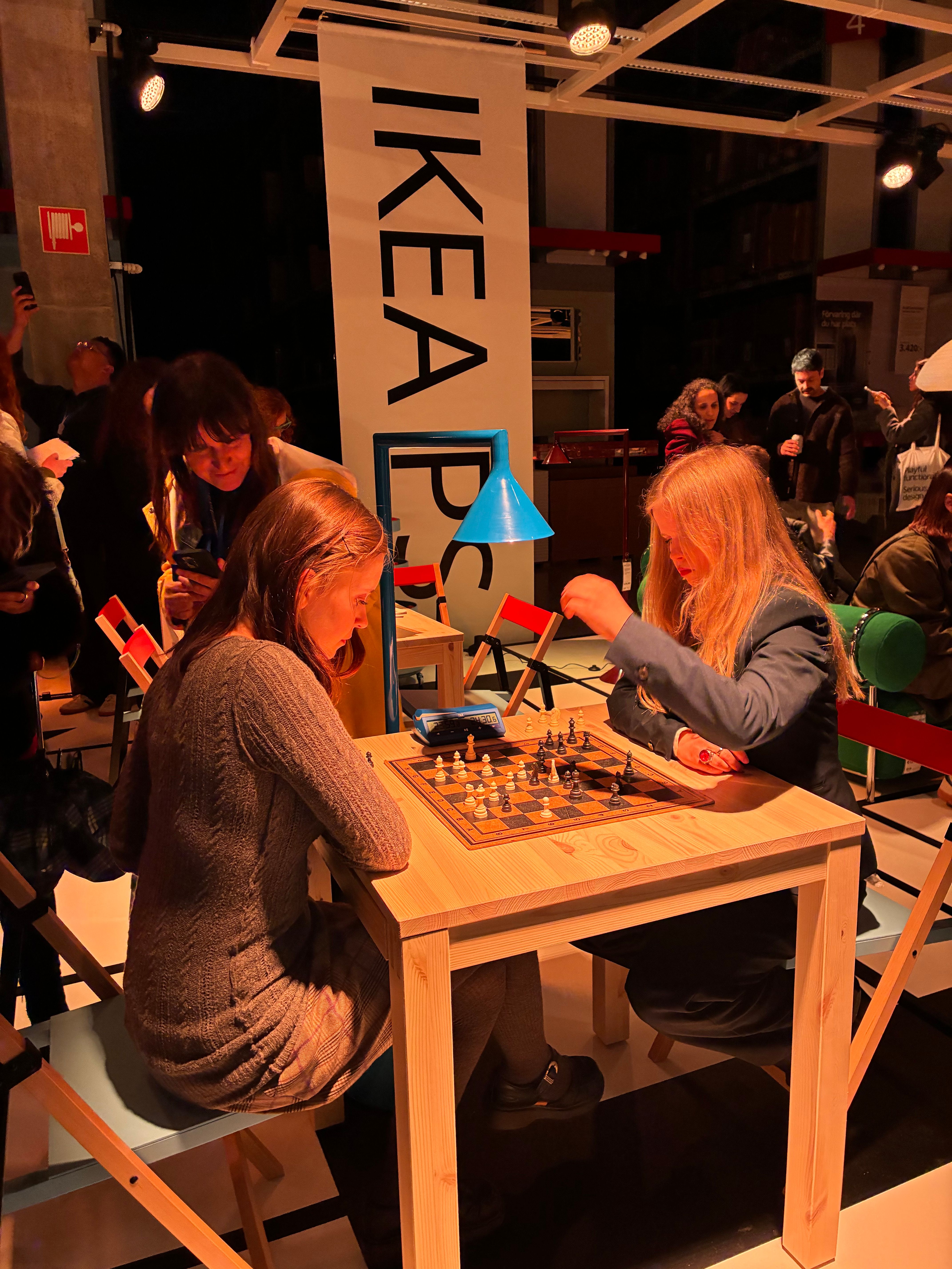 Two players at a chess match on a pine table beneath an IKEA PS banner and blue cone lamp at Democratic Design Days