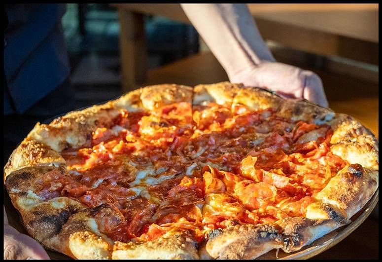 Hands lift a whole wood-fired pizza with a blistered charred crust and tomato-laden top at Osteria Antica
