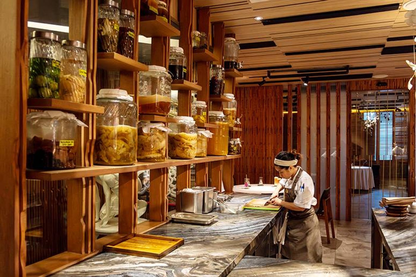 Wooden shelves of fermentation jars filled with fruits and chilies above a chef working at a marble counter