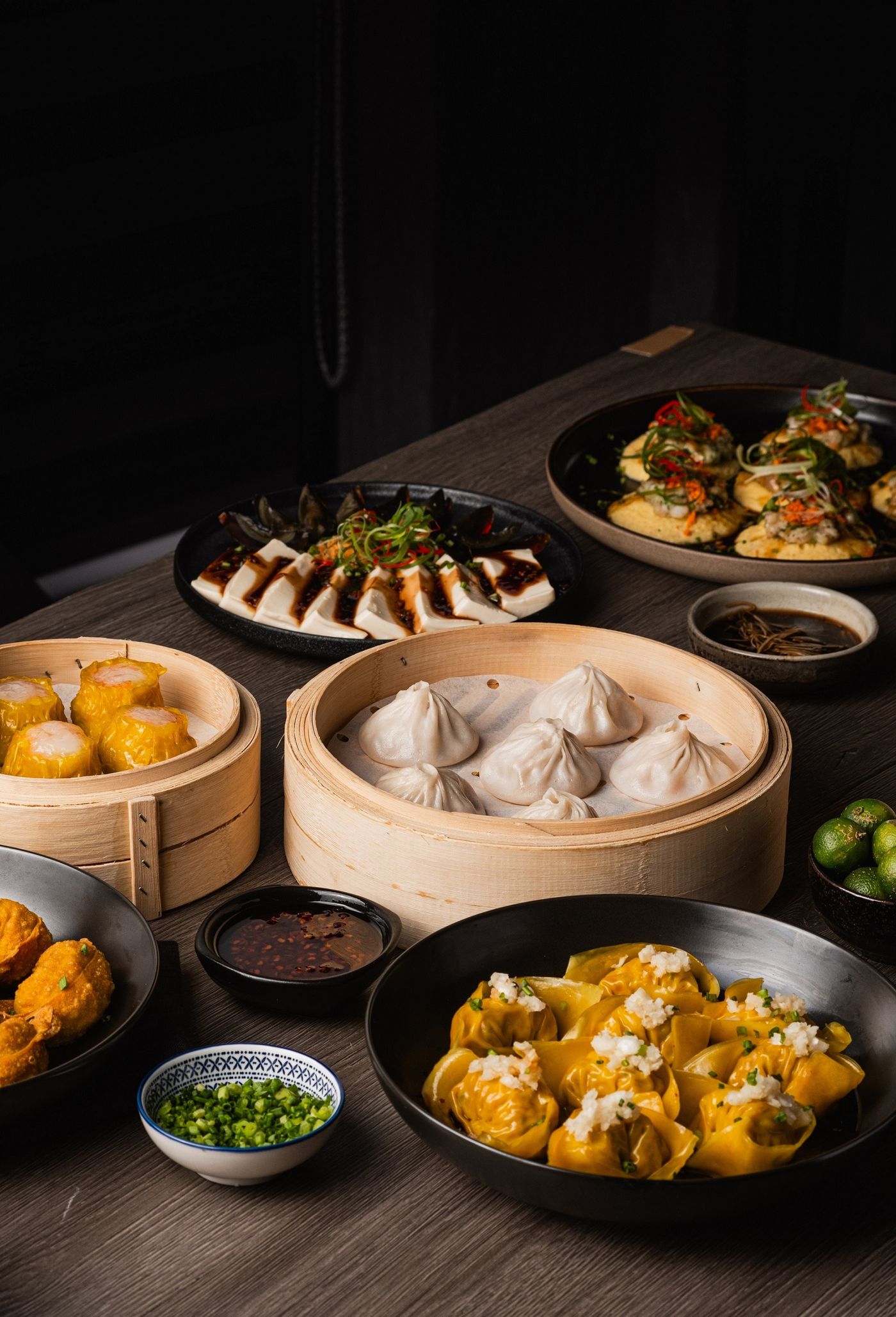 A wooden table spread filled with various Chinese dim sum dishes, including xiao long bao in bamboo steamers, pork siomai, and dumplings with dipping sauces