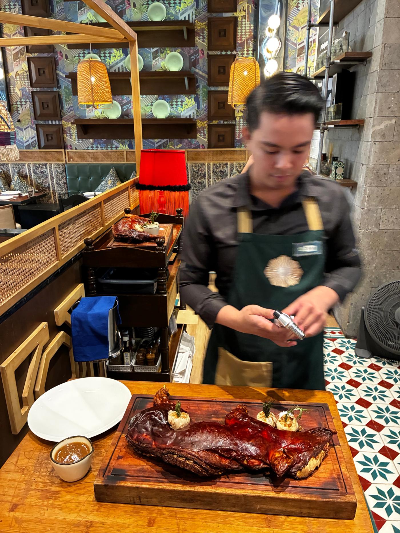 A server with a kitchen torch stands over a whole roast cochinillo on a wooden carving board, patterned tiles underfoot