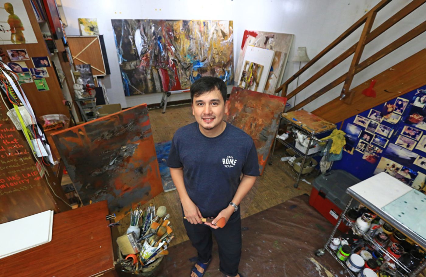 Martin Honasan stands in his studio surrounded by large abstract canvases, jars of brushes and a wall of pinned photographs