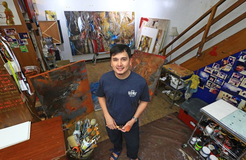 Martin Honasan stands in his studio surrounded by large abstract canvases, jars of brushes and a wall of pinned photographs