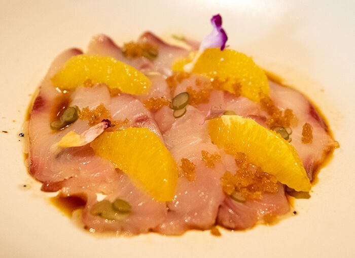 Slices of cured hamachi in a white bowl with citrus segments, orange roe and a dark soy dressing