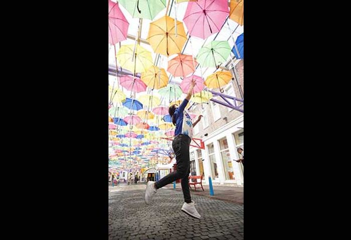 Hundreds of pastel umbrellas strung above a cobbled lane, with a visitor leaping mid-air, one hand raised to touch them