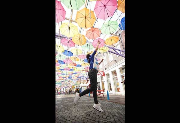 Hundreds of pastel umbrellas strung above a cobbled lane, with a visitor leaping mid-air, one hand raised to touch them