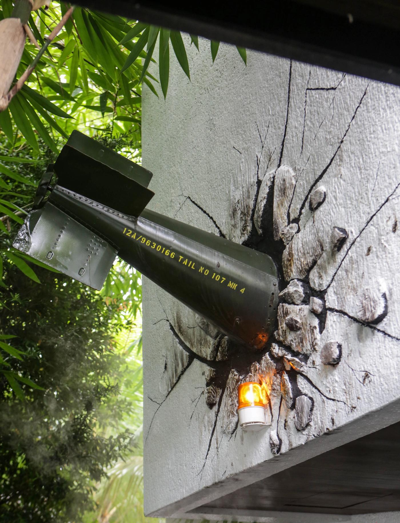 A green aerial bomb embedded nose-first in a cracked exterior wall, a small flame-shaped lamp glowing beneath it