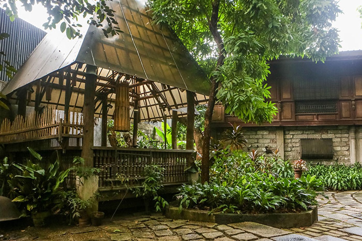 A bamboo and thatch bahay kubo built without nails standing in a rain-soaked garden, beside the capiz windows and stone wall of the main house